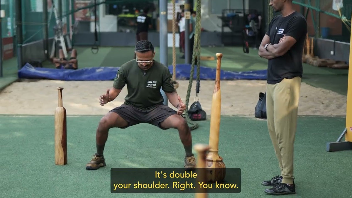 A Tagda Raho athlete in a deep squat holding wooden clubs at the training ground, with mudgars and a gada racked beside him